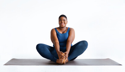 A woman sits on a yoga mat in a studio surrounded by a white background. She holds her feet while...