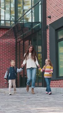 Vertical view of Caucasian mother walking with two children near brick building. Woman holding kids hands. Boy and girl wearing backpacks. Leaving school together. Family routine moment.
