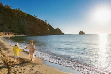 A woman in a white lace tunic and a young girl in a yellow shirt and sun hat walk hand-in-hand along a sandy shoreline at golden hour