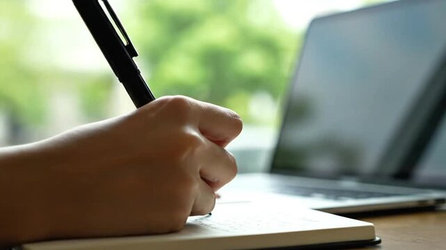 Close Up Hand Writing Notes in Notebook with Laptop in Background