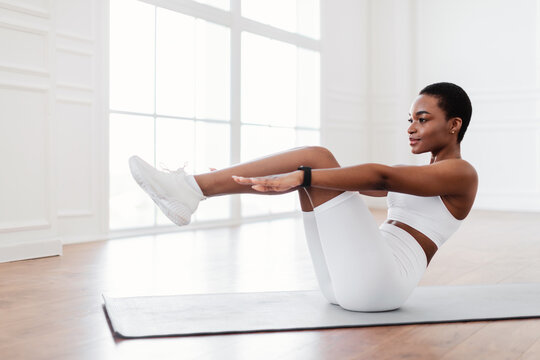 Abdomen Workout Concept. Strong confident young black female in white sportswear doing v-ups abs exercises on yoga mat in fitness club or at home in living room. Lady doing crunches with raised legs