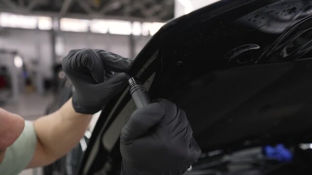 Automotive detailer in black gloves meticulously applying paint protection film on the edge of a black car's hood in a professional garage