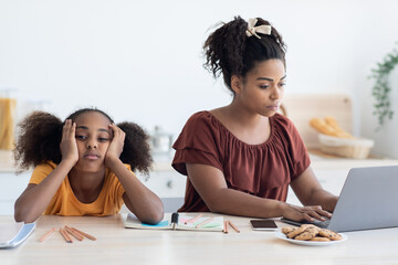 Obraz premium Black woman freelancer typing on laptop, working from home, bored daughter sitting by her, leaning on her hands, single mother does not paying attention to her kid teen girl, kitchen interior
