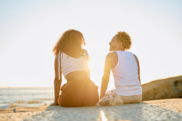 Diverse couple sitting on a concrete wall enjoying a romantic sunset over the ocean, embracing...