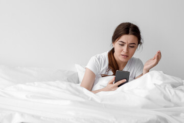 Confused shocked upset european young female received bad message, sits on white bed in bedroom interior, copy space. Human face expression, negative emotion, bad news reaction and device addiction