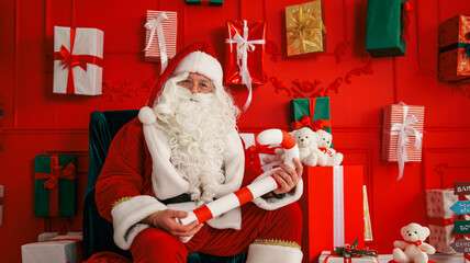 Santa Claus sitting in a chair surrounded by Christmas gifts and teddy bears