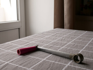 Metal tubular tool with red handle on a checkered tablecloth near a window, workshop still life.