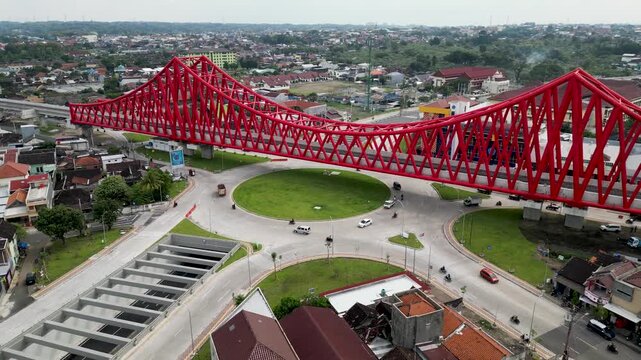 An aerial view of Simpang Joglo, Solo, featuring a striking red steel bridge above busy roads, buses, and cars, surrounded by dense urban neighborhoods and city life.