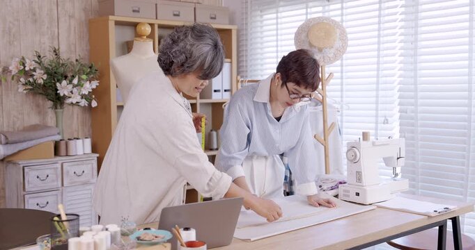 Senior asian woman leaning to assist daughter measuring fabric fashion boutique studio teamwork small business family designer sewing handmade textile entrepreneurship apparel garment pattern making