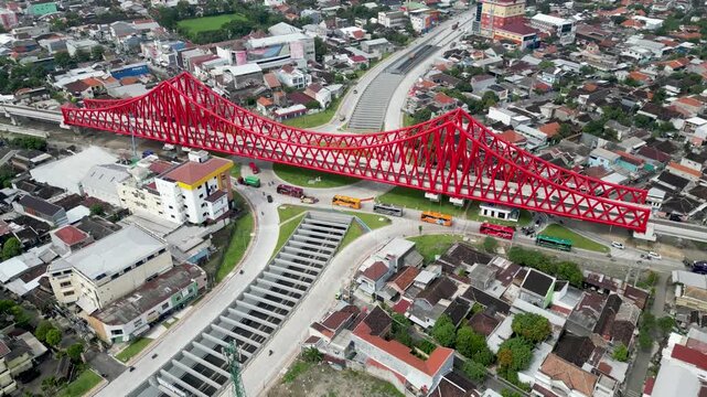 An aerial view of Simpang Joglo, Solo, featuring a striking red steel bridge above busy roads, buses, and cars, surrounded by dense urban neighborhoods and city life.