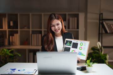 Asian businesswoman presenting charts and graphs on a clipboard during a remote video call, collaborating and analyzing financial data and statistics from her home office desk