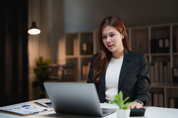Asian business woman working overtime in a dark office at night, using a laptop and calculator for financial tasks, concentrating on her diligent work