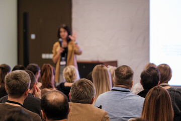 A confident presenter speaks to a seated audience in a conference room. Attendees listen attentively as a large screen glows in the background. Professional event atmosphere and learning environment.