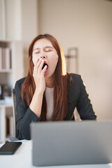 Young businesswoman covering her mouth while yawning, feeling sleepy and exhausted during prolonged work at her modern office with a laptop and phone on the desk