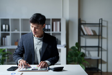 Young Asian businessman working late, reviewing financial reports and data charts on a clipboard, focused on business analysis and planning future strategies in a modern office setup