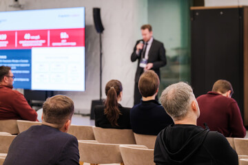 Audience sits in rows while speaker presents at conference. Large screen displays data and charts. Attentive participants in business attire engage with talk in corporate setting.