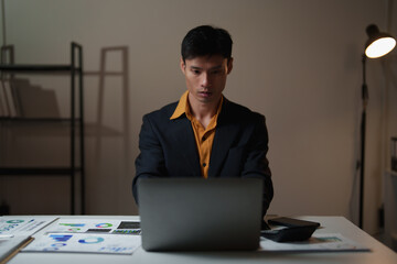 Asian businessman concentrating while typing on a laptop at his desk, surrounded by business documents and a calculator, illustrating dedication and evening work