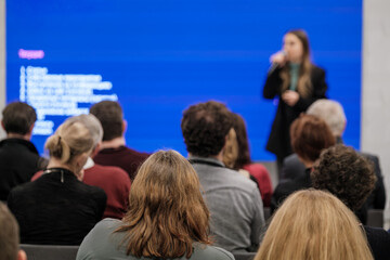 Audience seated facing speaker in conference room blue screen backdrop project slides professional presentation atmosphere.