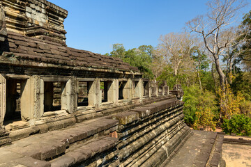 Fototapeta premium A row of open stone windows lines the elevated terrace of Baphuon temple in Angkor, Cambodia, under bright sunlight. Weathered sandstone and intricate carvings contrast with the surrounding trees and