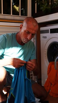 Mature man in casual clothes sorting blue sportswear by a washing machine, carrying out everyday laundry chores in his apartment as part of a morning home routine