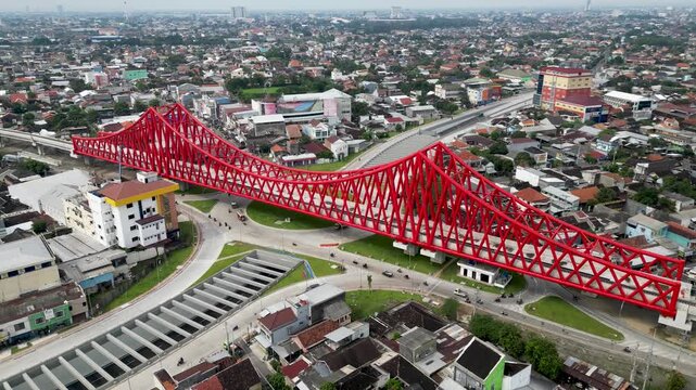 An aerial view of Simpang Joglo, Solo, featuring a striking red steel bridge above busy roads, buses, and cars, surrounded by dense urban neighborhoods and city life.