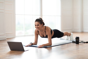 Fototapeta premium Bodyweight Workout Routine. Cheerful Fit Woman Standing In Plank Position On Yoga Mat At Fitness Studio Or Home Looking At Computer Screen Watching Online Tutorial Making Video Call With Trainer