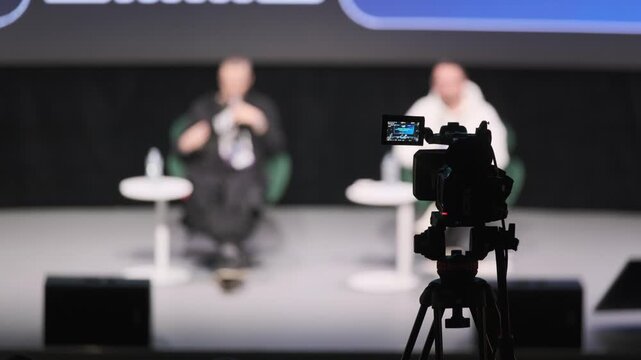 Blurred stage interview scene with two speakers on panel captured by professional camcorder in studio setup, showcasing modern media production and focused discussion