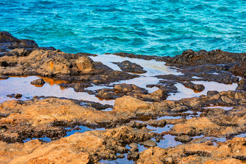 Lava rock with tide pools. Dark volcanic rocks line shore, forming tide pools that mirror sky. Turquoise waves roll gently in background, contrasting rugged foreground with serene ocean