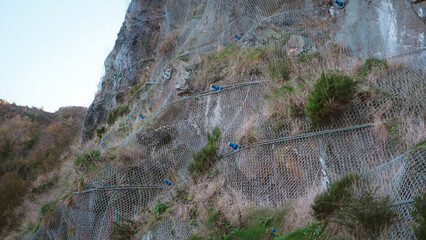 A low-angle detailed view of a steep rocky cliff face reinforced with a heavy-duty steel wire mesh netting designed to prevent rockfalls and landslides.