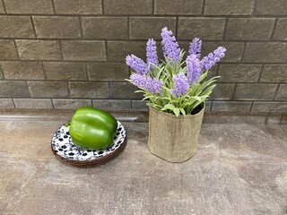 Green bell pepper on decorative plate beside a burlap vase filled with lavender flowers, set against a textured wall, creating a vibrant kitchen atmosphere