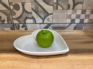 Green apple resting on a white heart-shaped plate, placed on a wooden surface, with a patterned tile background, creating a charming and inviting kitchen atmosphere