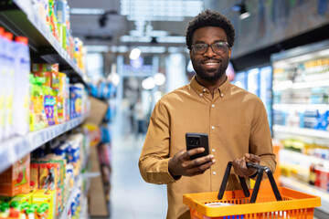 Smiling african american man using a mobile phone and holding a shopping basket while navigating the aisles of a modern supermarket, managing his grocery list