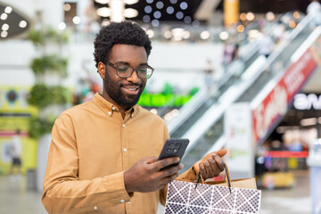 Happy young man wearing glasses holding a branded shopping bag, smiling while checking his smartphone for notifications, messages, or online shopping deals in a modern shopping mall