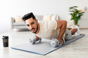 Fototapeta premium Stay at home fitness. Strong young Arab man doing whole body workout, standing in plank pose, using dumbbells indoors. Fit Eastern guy strengthening abs muscles in living room
