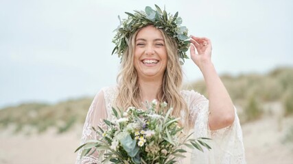 Smiling young bride in a bohemian lace dress holding a flower bouquet. Happy woman wearing a green leaf crown laughing on a beach with sand dunes. Bridal portrait in nature