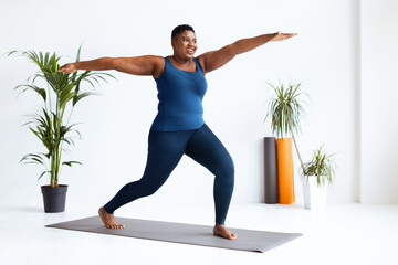 A woman stands in a yoga pose on a mat in a spacious studio. She has a focused expression and is...
