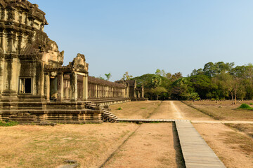 Fototapeta premium Side view of the ancient sandstone gallery at Angkor Vat, Cambodia, with warm sunlight highlighting intricate Khmer carvings and balustered windows. The temple structure opens onto a dry grassy