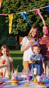 Outdoor backyard birthday party with colorful table, parents and children smiling under bunting flags, cake, balloons, juice and snacks on sunny summer afternoon with warm festive atmosphere