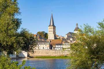Fototapeta premium Distinctive abbey tower rises above the historic rooftops of La Charite sur Loire, framed by leafy trees and the calm Loire river under a clear blue sky. The scene highlights the medieval architecture