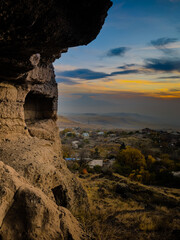 A rugged cave opening frames a picturesque landscape of a distant town and rolling hills under a clear sky. Sunlight highlights the warm tones of the rock formations surrounding the entrance. High