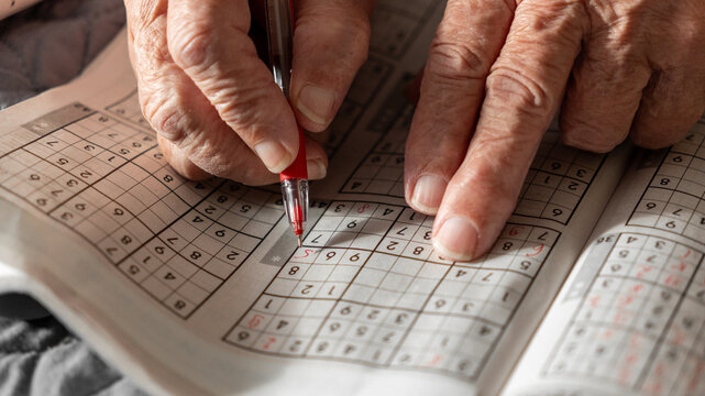 old woman, pensioner, grandmother solves Sudoku crosswords and writes numbers in a journal with a red pen. entertainment for pensioners, leisure for the elderly, brain activity, thinking,mental health