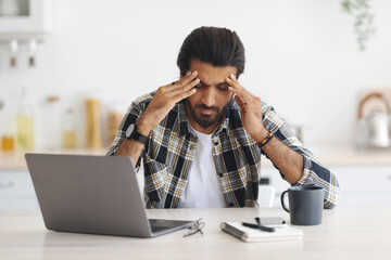 Exhausted indian man sitting in front of laptop at home, touching his head, suffering from burnout...