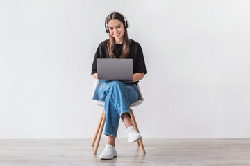 Cheery Caucasian woman in headphones having online video call on laptop, sitting on chair against...