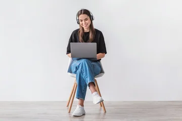 Fotobehang Diamanten Cheery Caucasian woman in headphones having online video call on laptop, sitting on chair against white wall, full length. Millennial lady participating in webinar, communicating remotely on internet  © Prostock-studio