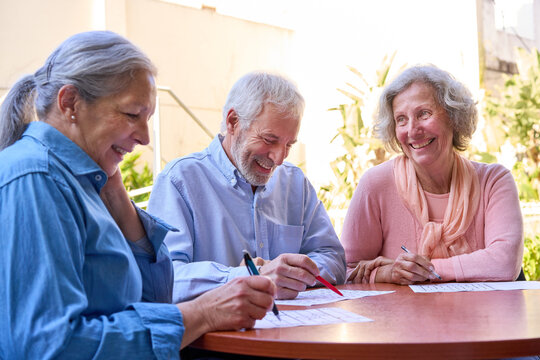 Senior People Doing Cognitive Rehabilitation Exercises Outdoors At Rehab Center Writing On Paper With Pens Smiling And Laughing Together In The Sunlight