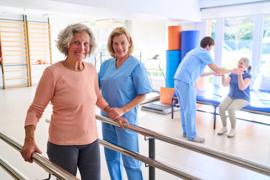 Senior Woman Walking With Parallel Bars Assisted By Physiotherapist In Rehab Center During Rehabilitation Session Indoors