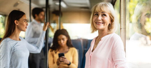 Public Transportation Concept. Portrait of smiling senior lady standing in bus, leaning looking out of window, going home, banner, panorama. Group of diverse people passengers in blurred background © Prostock-studio