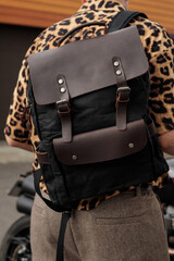 A classic leather backpack slung over the shoulder of a guy wearing a leopard print shirt.
