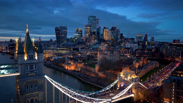 Aerial view of the Tower of London with the illuminated city skyscrapers behind and the Tower Bridge in front during evening time