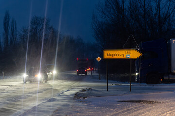 Night winter road Magdeburg heavy snowfall cars truck driving icy junction illuminated headlights streetlight. Direction sign low visibility snow covered asphalt traffic danger transport challenge